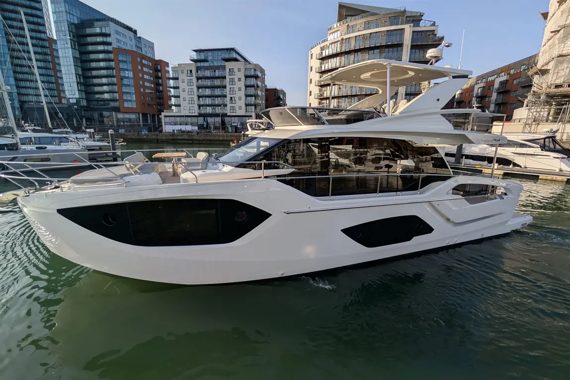 A sleek white yacht docked in a marina, surrounded by modern buildings and other boats, reflecting in the water.